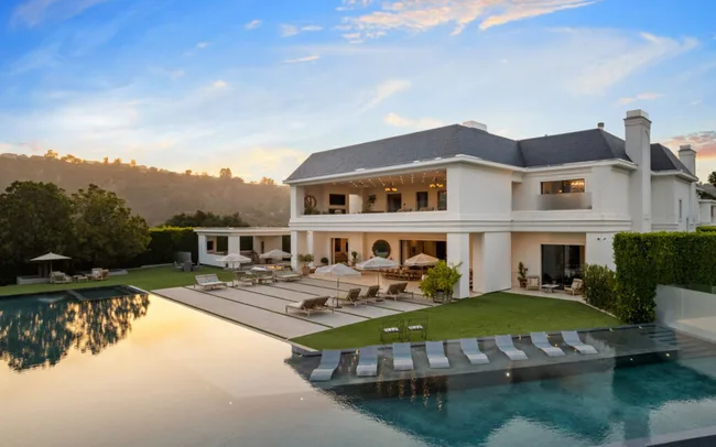 Elegant luxury home with a pool, patio, and lawn set against a backdrop of hills at sunset.