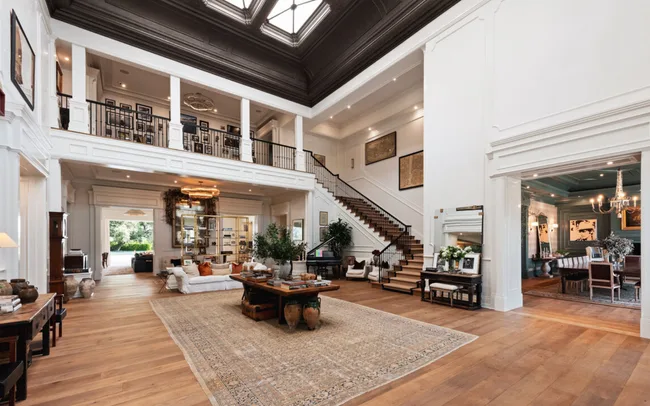 Spacious, elegant living room with wooden floors, large rug, grand staircase, and a dining area visible through an open doorway.