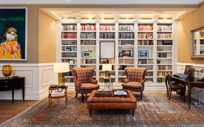 Cozy library with plaid chairs, bookshelves, a leather ottoman, and a vibrant painting on the left wall.