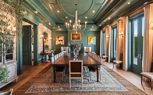 Elegant dining room with long wooden table, chandelier, green walls, large windows, and contemporary art.