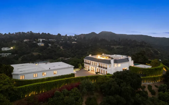 Aerial view at dusk of a large, elegant white mansion surrounded by lush greenery and rolling hills.