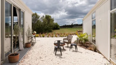 Outdoor seating on a gravel patio between modern buildings, with potted plants and grassy field under a cloudy sky.