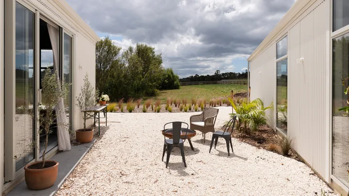 Outdoor seating on a gravel patio between modern buildings, with potted plants and grassy field under a cloudy sky.