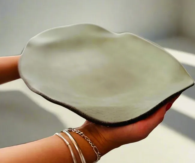 A person holding an abstract, leaf-shaped ceramic plate in neutral tones.