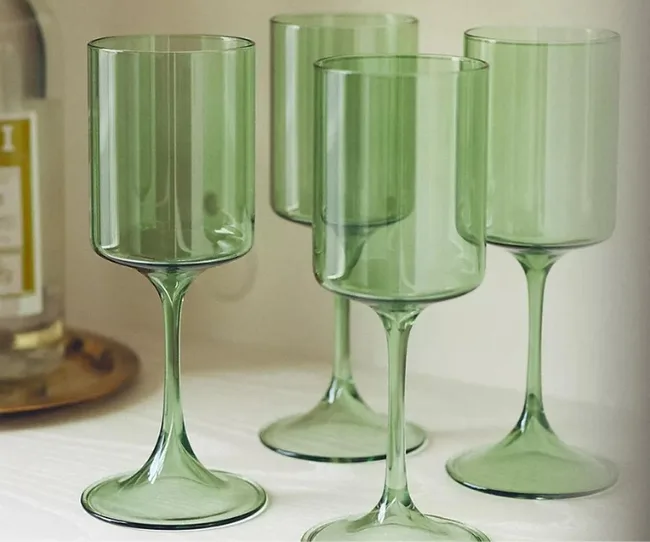 Four translucent green wine glasses on a white surface with a blurred bottle in the background.