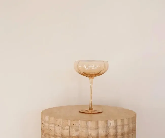 A clear glass coupe sits elegantly on a textured, round stone pedestal against a soft beige background.
