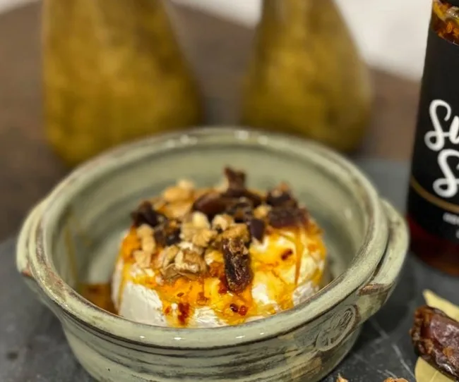 Baked cheese with nuts and drizzle in a ceramic dish, with pears and a bottle in the background.