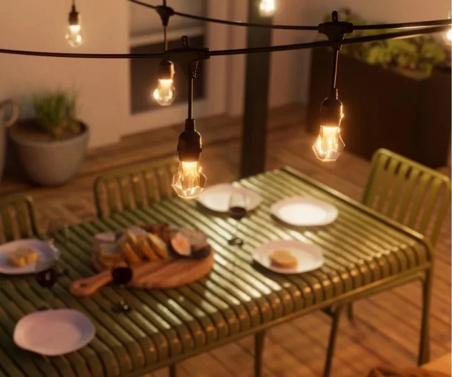 Outdoor dining setup with string lights, plates, and a wooden board with food on a green table.