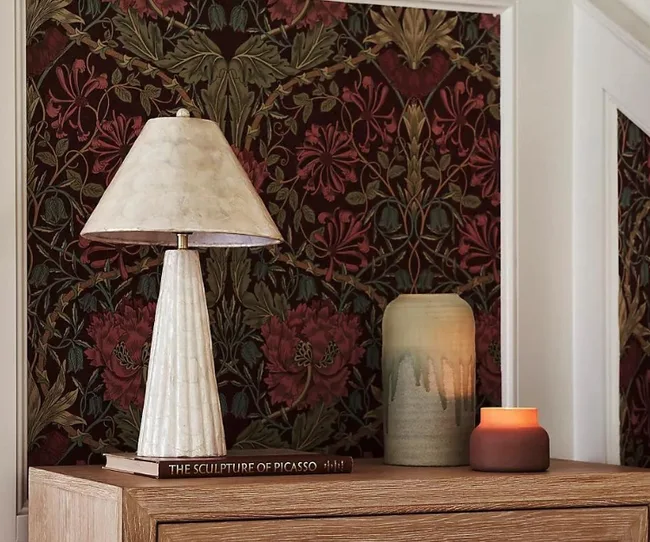 A table lamp, a vase, and a small candle on a wooden surface with floral wallpaper background.