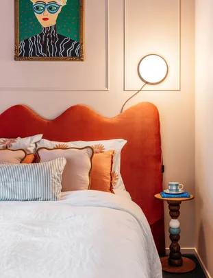 Bed with orange headboard, pillows, bedside table with book and mug; artwork of a person with glasses on the wall.