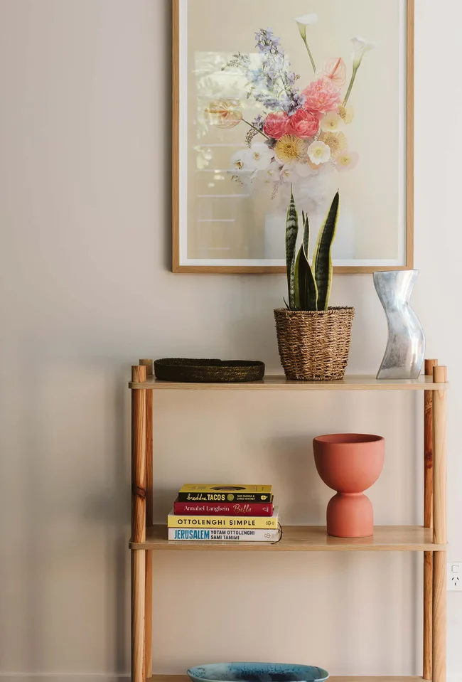 Shelf with decor items, including a plant, books, and a vase, beneath a framed floral artwork against a light wall.