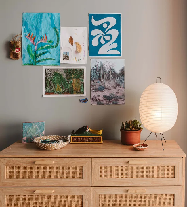Artwork and decor on a wall above a dresser with a basket, plant, and glowing lamp.