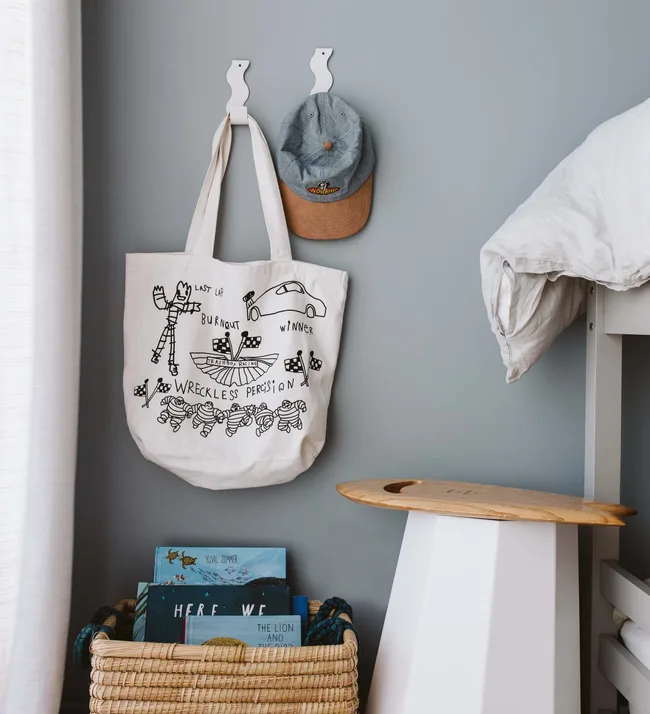Canvas tote with race car drawings and text. Gray cap hangs on a hook. Rattan basket with books beneath.