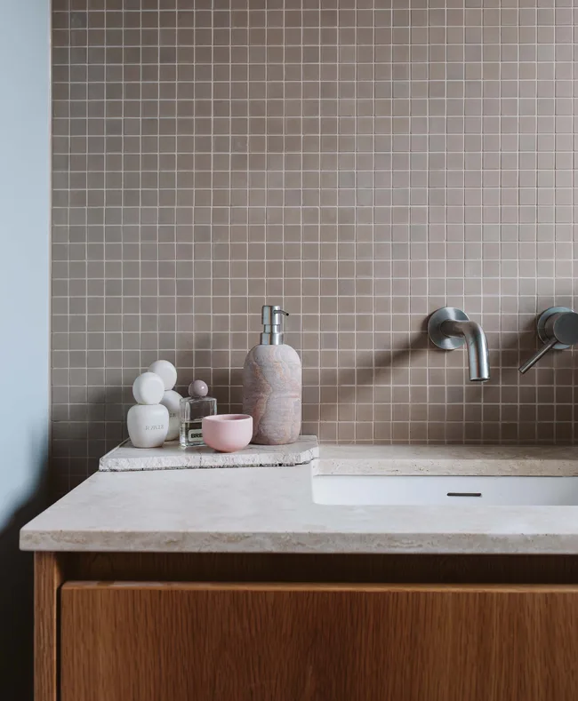 Bathroom sink with beige tiles, marble soap dispenser, toiletries, and wall-mounted faucet on a wooden vanity.
