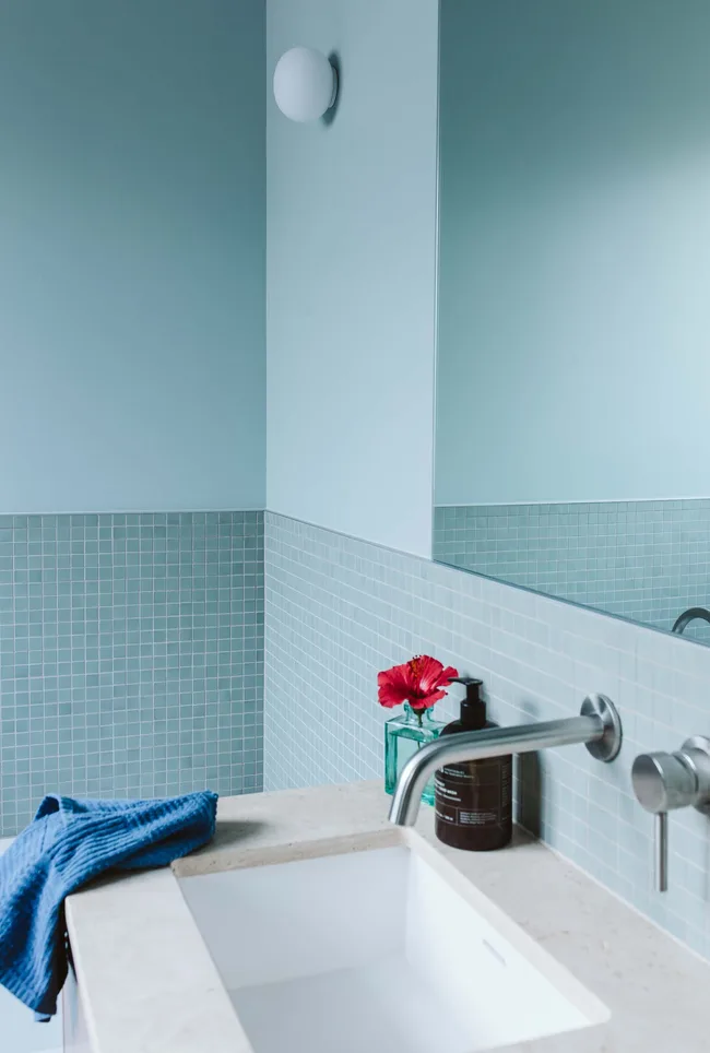 Modern bathroom with blue walls, square tiles, a silver faucet, a red flower in a vase, and a soap dispenser.