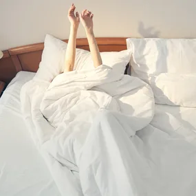 Person stretching under white bedding in a sunlit room, with only arms visible.