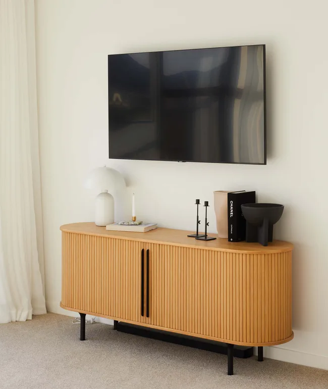 Modern living room with a mounted TV, wooden sideboard, decorative items, and a standing lamp.