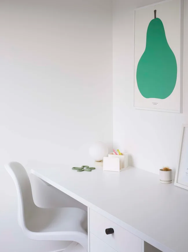 Minimalist white desk setup with a chair, green decor, and pear art on the wall.