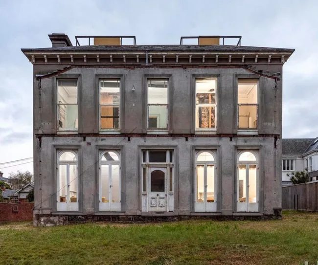 Abandoned two-story mansion with large windows and overgrown lawn, appearing aged and uninhabited.