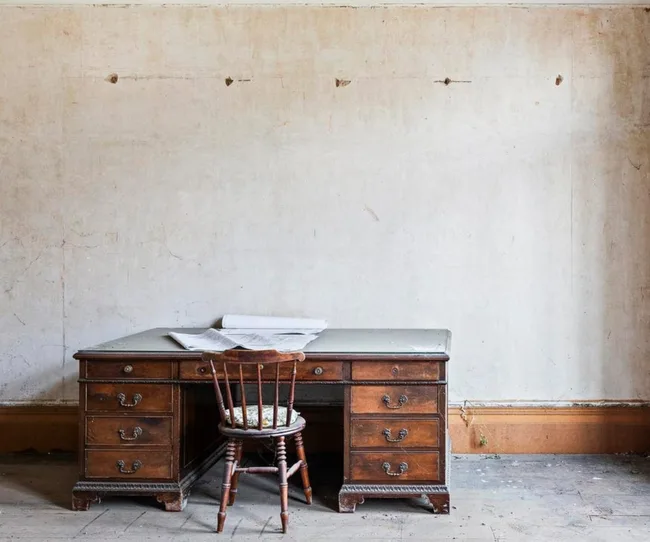 Old wooden desk and chair in an empty room with peeling paint and rolled papers on the desk.