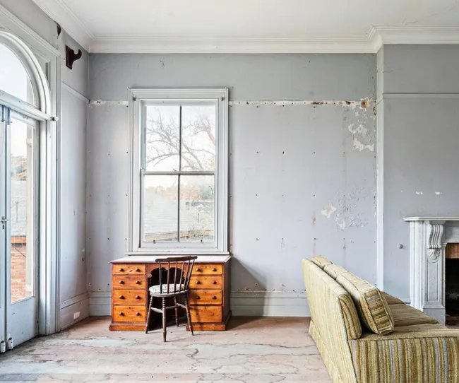 A room with a wooden desk, chair, striped sofa, and peeling paint walls beside a large window.