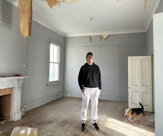 Man in casual attire stands in room with peeling ceiling; a small dog is nearby.