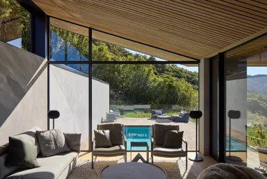 Modern living room with large windows, views of a deck and pool, surrounded by greenery and hills.