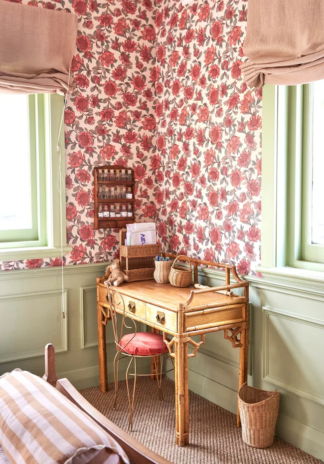 A vintage wooden desk in a floral wallpapered room with a wicker basket, chair, and window with beige curtains.