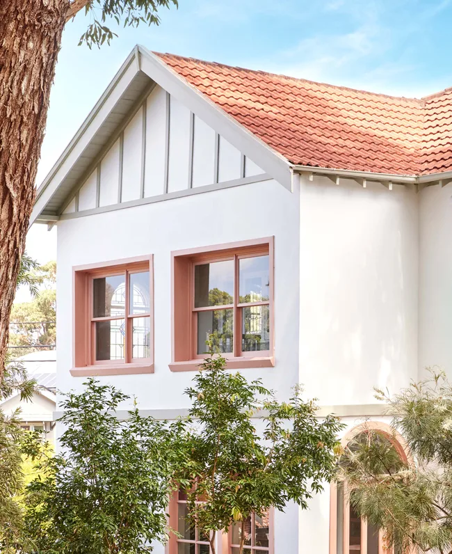 Red-tiled roof and white walls of a house with pink-framed windows, surrounded by trees under a blue sky.