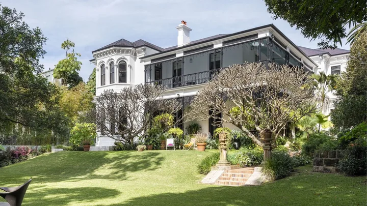 Elegant white mansion with large windows and wraparound balcony, surrounded by lush gardens and trees under a blue sky.
