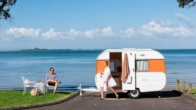 A woman in white dress walks near an orange caravan by the sea, while a man sits on a chair with drinks.