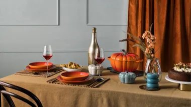 Autumn-themed dining table set with orange dishes, wine glasses, flowers, and a pumpkin-shaped pot.