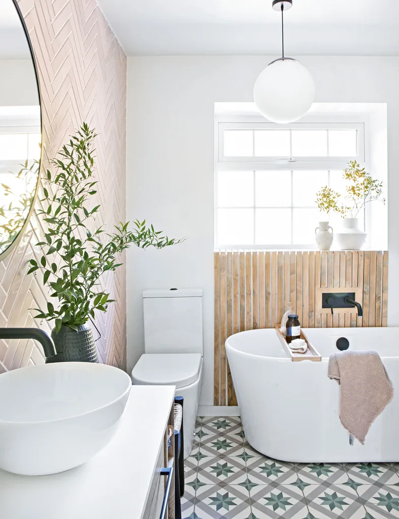 A bathroom with a toilet, bath and vanity, pattern tiled floors and pink herringbone tiled walls.