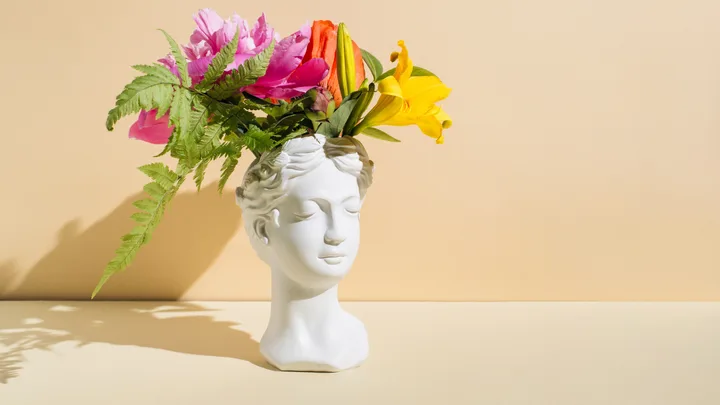 A vase shaped like a woman's head with a bouquet of flowers, against a beige background.