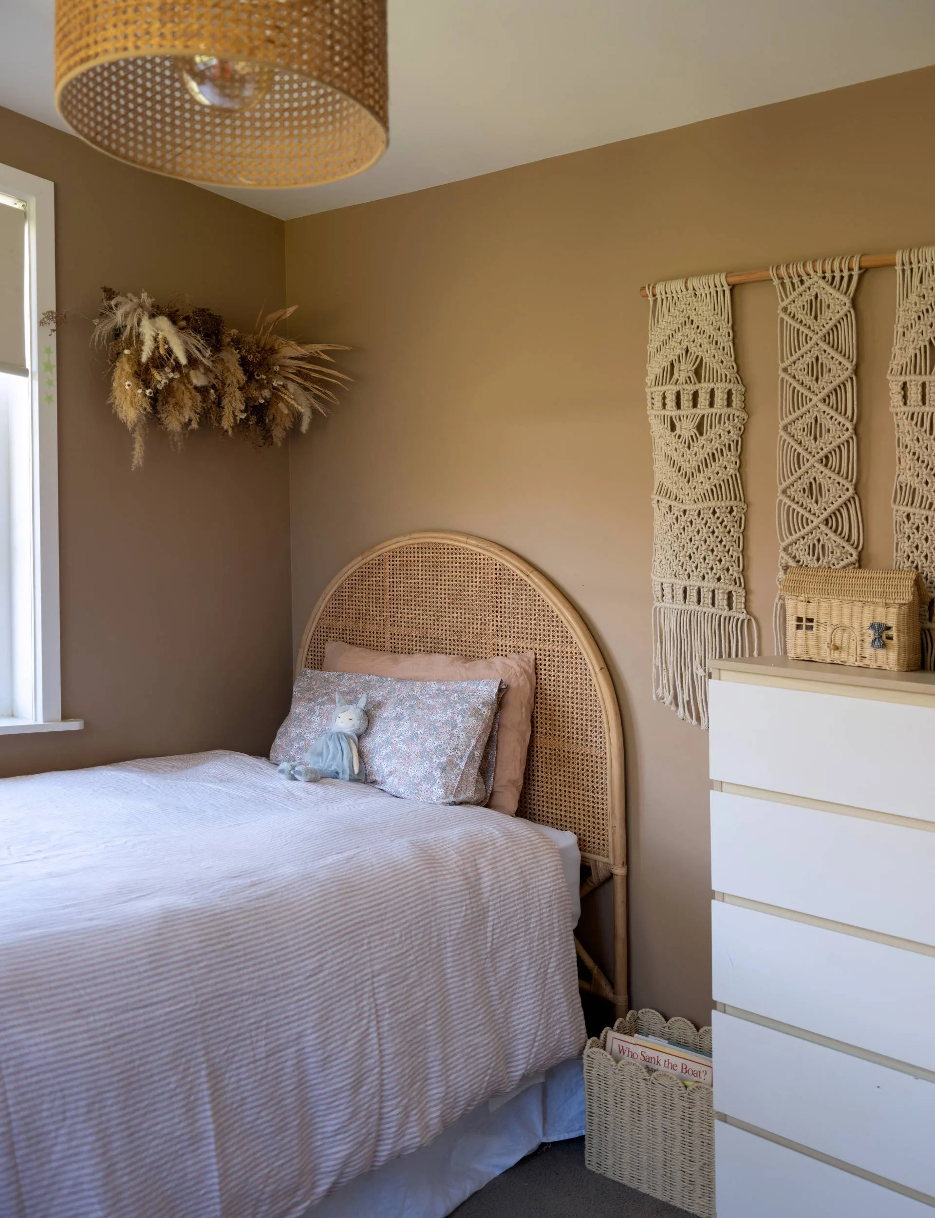 A brown bedroom with a single bed in the corner, a pink and white striped duvet cover over it and natural string woven art on the walls