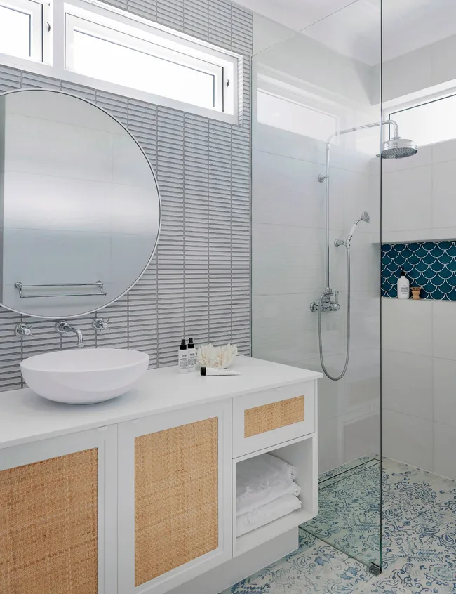 The Hamptons-style bathroom has grey-blue tiles on both the walls and floor, with a large rod mirror above a rattan and white vanity