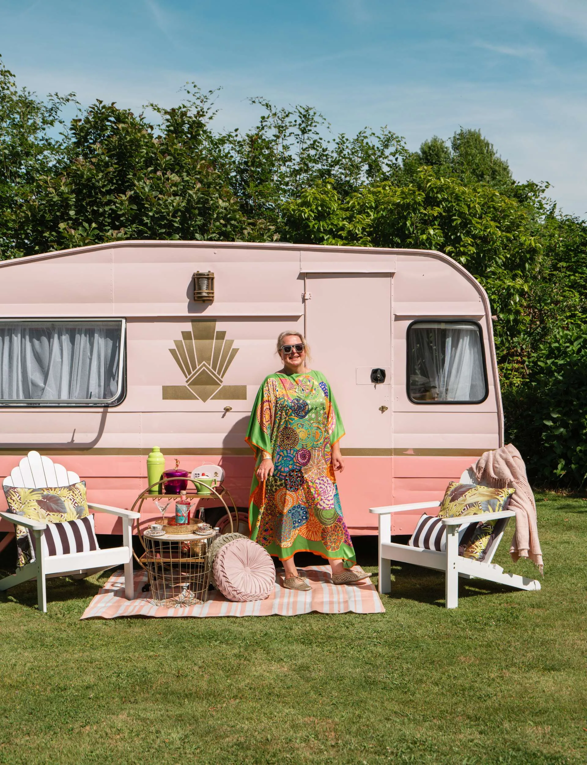 woman standing outside of pink caravan surrouded by a hedge and gress