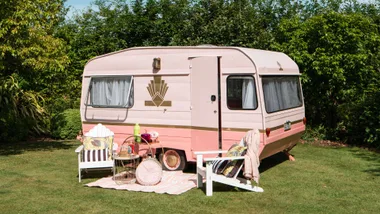 Pink caravan with chairs sitting out the front with grass and a huedge surrounding it.