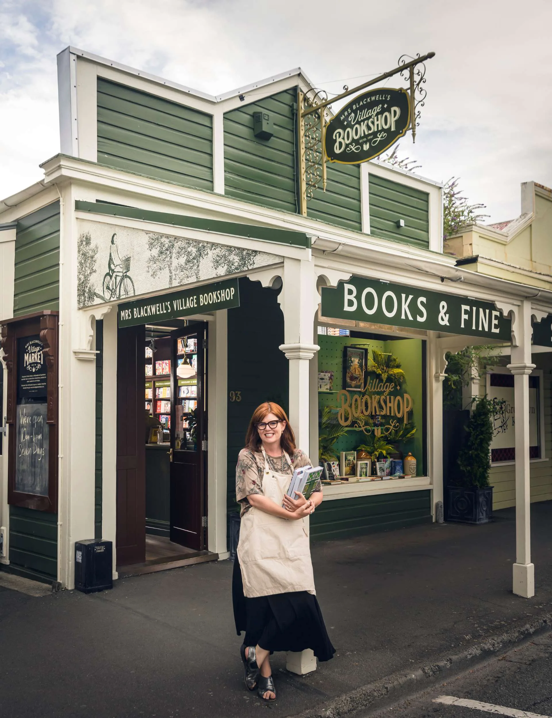 woman standing outside of green bookshop while holding some books.