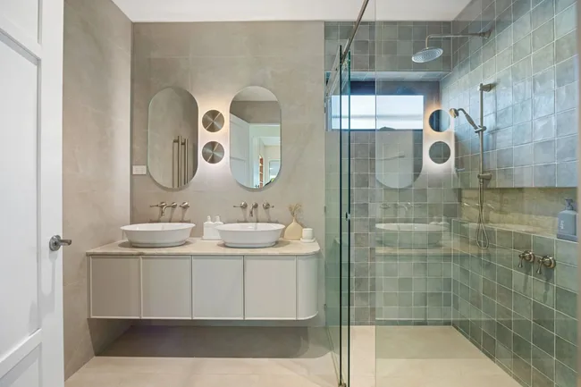 Modern bathroom with dual sinks, oval mirrors, and a glass-enclosed shower on tiled walls.