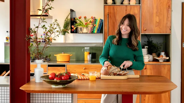 A woman wearing a green jumper cutting bread in her kitchen.
