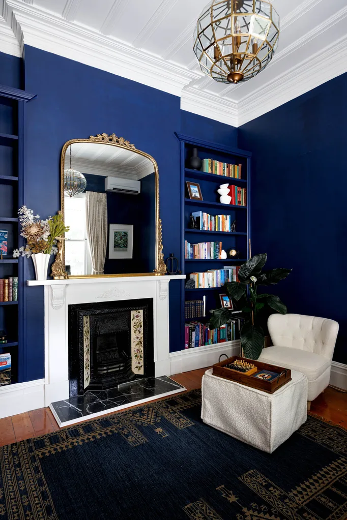 A home library space with royal blue walls and a white ceiling. An ornate mirror sits above the fireplace and a white armchair is in the corner.