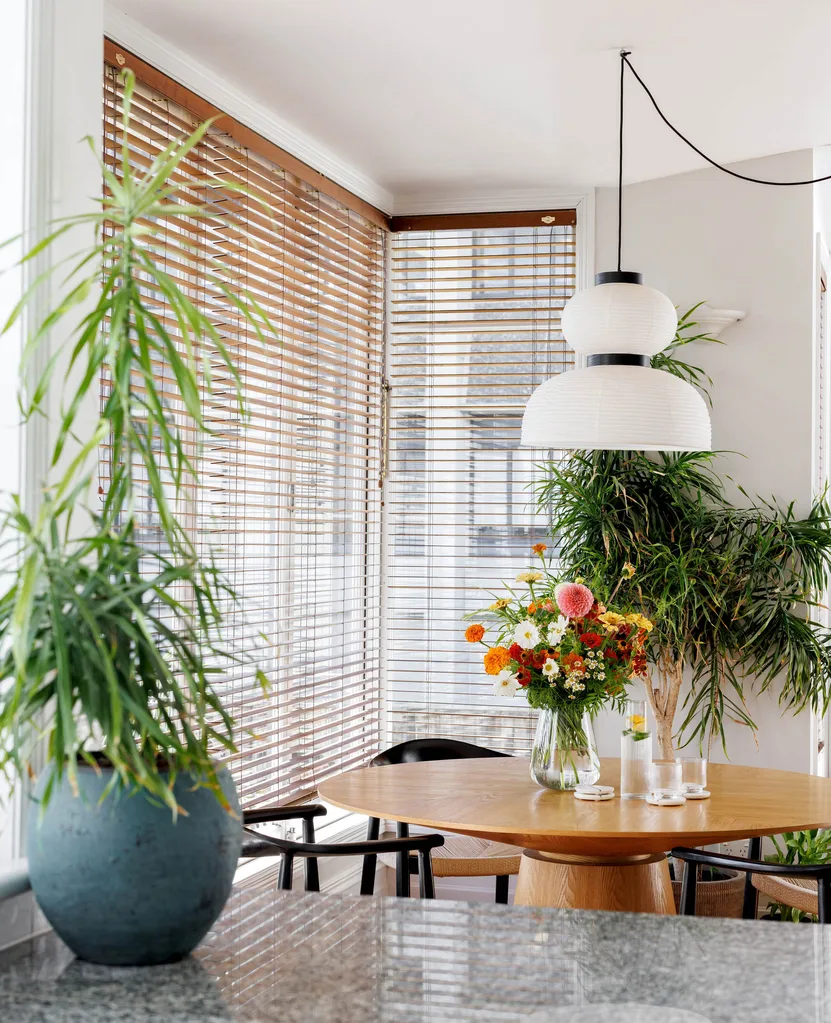 Looking into a dining room with a round dining table, topped with a bouquet of flowers. Blinds cover the windows and a white pendant light hangs from the ceiling.