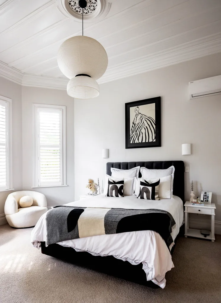 A main bedroom with black and white bedding, a print on the wall, a white pendant hanging from the ceiling and a white armchair in the corner.