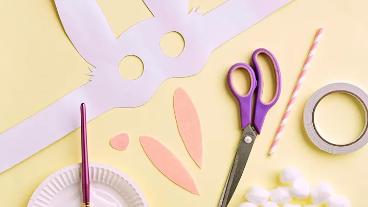 A bunny mask, scissors and a paint brush on a yellow background.