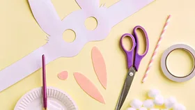 A bunny mask, scissors and a paint brush on a yellow background.