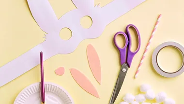 A bunny mask, scissors and a paint brush on a yellow background.