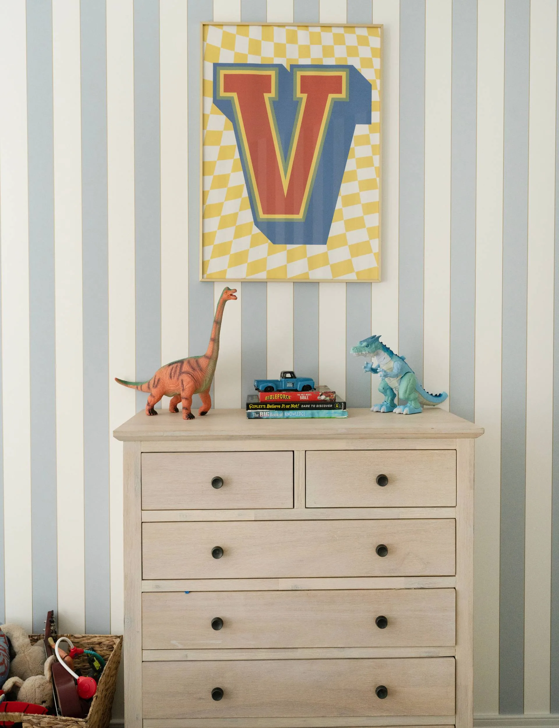 A wooden chest of drawers against a vertical blue and white striped wallpaper wall, with framed art above