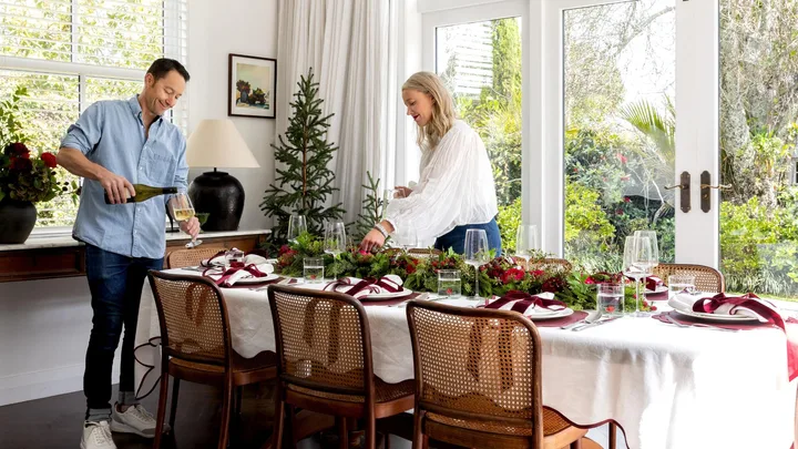 A man pours wine while a woman arranges a festive table set for six in a bright dining room decorated with greenery and red accents.