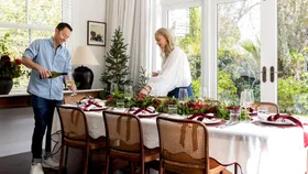 A man pours wine while a woman arranges a festive table set for six in a bright dining room decorated with greenery and red accents.